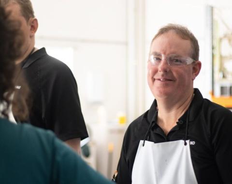 A hoto of a person at work smiling at the camera wearing goggles and an apron.